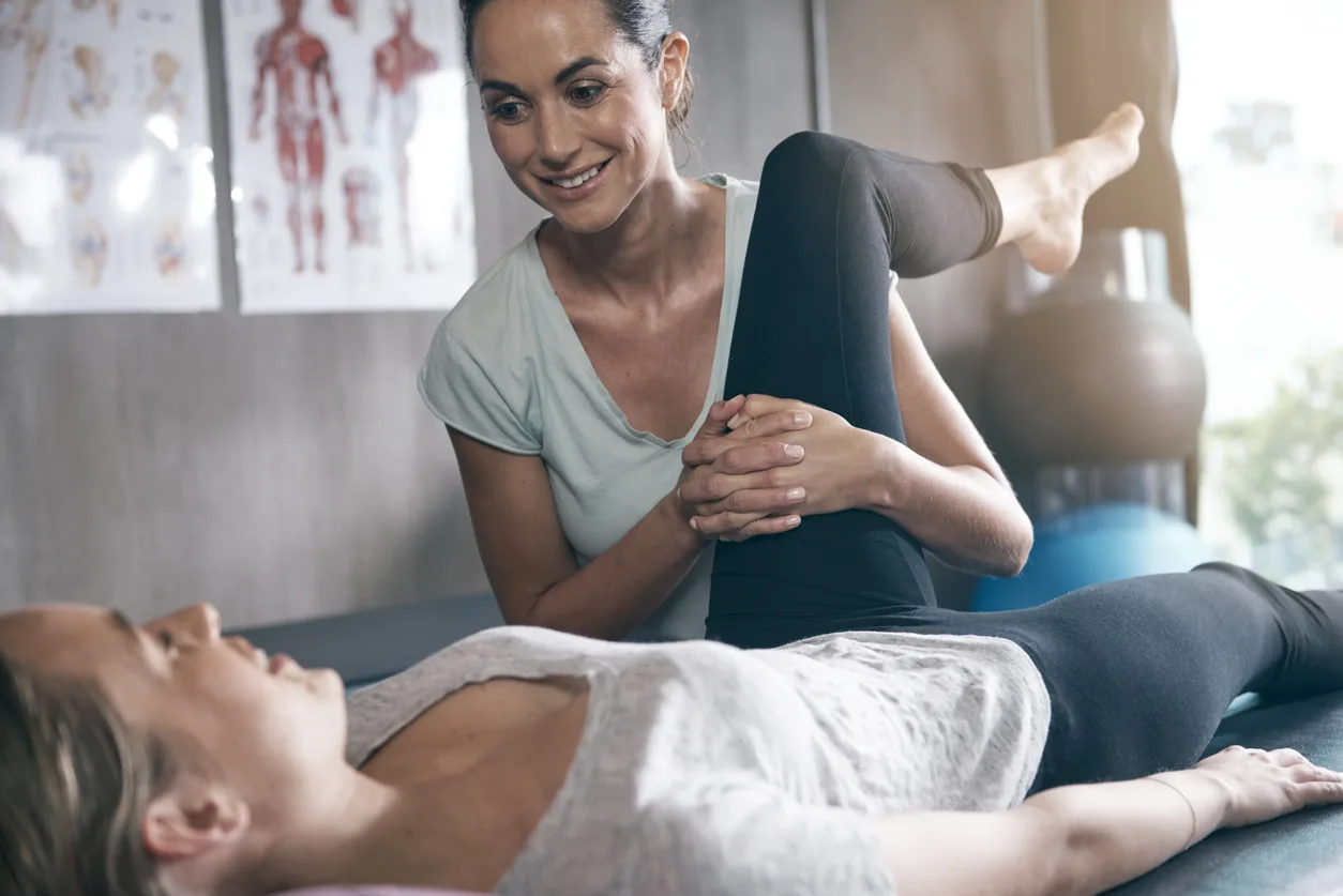 A woman is stretching her leg while another person lies on the ground.